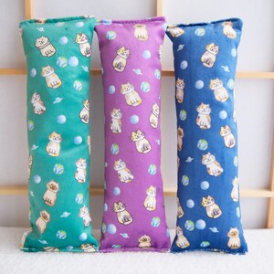 Puede incluir: Tres juguetes para gatos de tela verde, morado y azul. Cada juguete presenta un patrón repetitivo de gatos de dibujos animados y pelotas.