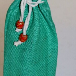 May include: A green burlap drawstring bag with a white cord and red wooden beads. The bag is lined with a gold fabric.