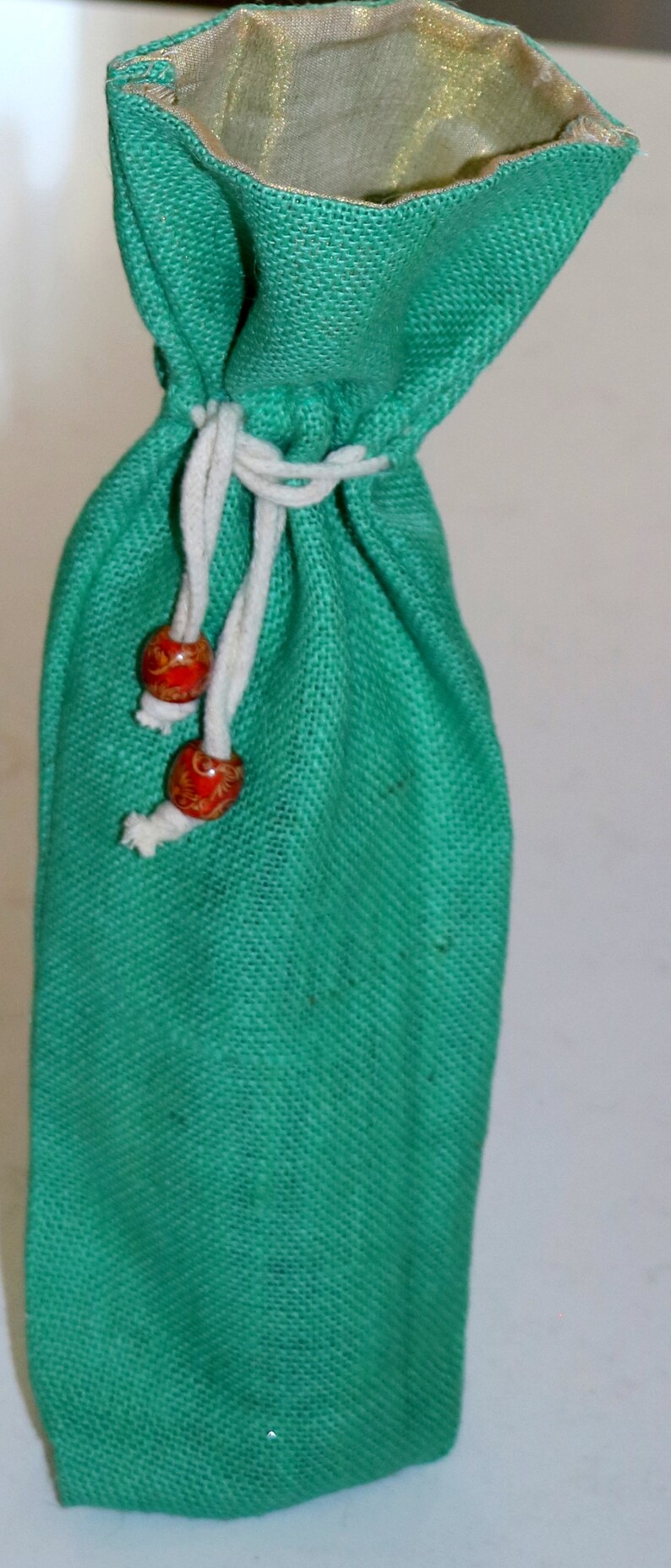 May include: A green burlap drawstring bag with a white cord and red wooden beads. The bag is lined with a gold fabric.