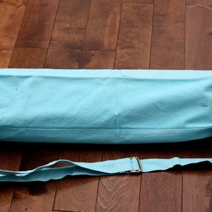 May include: A light blue yoga mat bag with a drawstring closure and an adjustable shoulder strap. The bag is made of canvas and has a simple design.