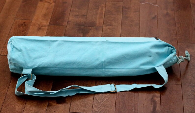 May include: A light blue yoga mat bag with a drawstring closure and an adjustable shoulder strap. The bag is made of canvas and has a simple design.