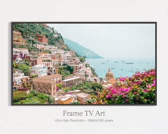 May include: A framed photograph of a colorful Italian village nestled on a hillside overlooking the ocean. The village is filled with buildings in shades of yellow, orange, red, and pink, and there are lush green trees and flowers throughout. The ocean is a bright blue, and there are boats in the distance. The image is framed in a black frame.