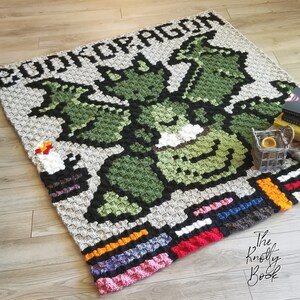 May include: A crochet blanket featuring a green dragon with the text "Book of Dragon" in black. The blanket has a colorful border with red, orange, yellow, black, blue, and pink.