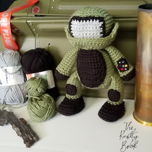 Könnte beinhalten: Ein grünes und braunes gehäkeltes Plüschtier in Form eines Roboters. Der Roboter hat einen großen Kopf mit einem weißen Visier und einen braunen Körper mit schwarzen Akzenten. Das Spielzeug sitzt auf einer weißen Oberfläche.
