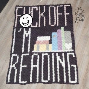 Może przedstawiać: Czarno-biały koc wykonany na szydełku z napisem "F*CK OFF I'M READING" i stosem kolorowych książek na półce. Koc wykonany jest z fakturowanym ściegiem i ma białą obwódkę.
