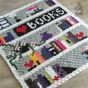 May include: A crochet blanket with a gray and white background featuring a bookshelf with colorful books and the text "I LOVE BOOKS".