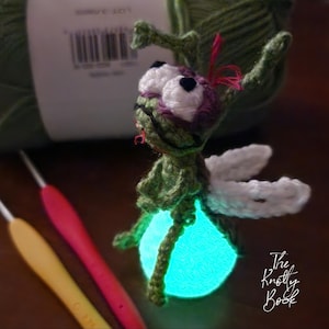 Può includere: Una lucciola all'uncinetto fatta a mano con una pancia turchese brillante. La lucciola ha ali bianche, grandi occhi bianchi e un corpo verde. Il logo The Knottly Book si trova nell'angolo in basso a destra. Uncinetti e filato verde sono sullo sfondo.