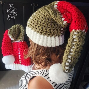 May include: A green, white, and red crocheted hat with a long, pointed top. The hat has a white pom-pom on the end of the top. The hat is being worn by a person with brown hair.