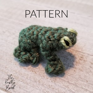 May include: A green crocheted frog with yellow eyes and a white background. The word "PATTERN" is written in black above the frog.