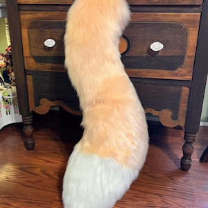 May include: A plush, curved tail with a peach-colored top and a white tip. The faux fur tail is displayed against a dark wooden dresser with white drawer pulls, and a wooden floor.