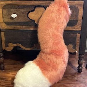 May include: A plush, orange and white faux fur tail. The tail is a vibrant orange color with a white tip. The tail is curved and fluffy, and appears to be made of soft material. The tail is standing upright on a wooden surface.