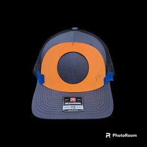 Könnte beinhalten: Ein grauer und schwarzer Trucker-Hut mit einem orangenen Kreis und einem schwarzen Loch in der Mitte. Der Hut hat ein Etikett mit der Aufschrift "EST. 1970 RICHARDSON. ORIGINAL 112 TRUCKER ADJUSTABLE".