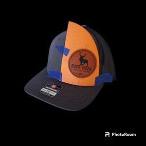 Puede incluir: Una gorra de camionero negra y naranja con un parche de cuero marrón que presenta una silueta de un ciervo y el texto "Buck Fiden I Will Not Comply". La gorra es ajustable y tiene una parte posterior de malla.