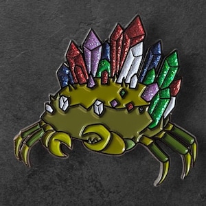 May include: A crab-shaped enamel pin with a crown of colorful crystals. The crab is olive green with yellow claws and legs. The crystals are red, purple, green, and white, with glitter accents. The pin is set against a dark gray background.