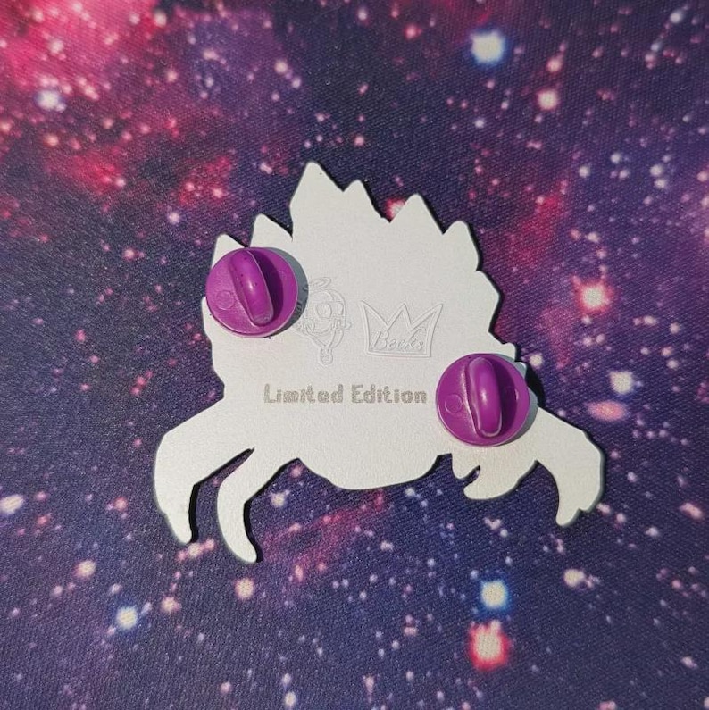 K&ouml;nnte beinhalten: Ein wei&szlig;er Emaille-Ansteckpin mit einem stilisierten Krabben-Design, mit zwei lila Anstecknadeln. Der Pin tr&auml;gt den Text "Limited Edition" und ein kleines Kronenlogo. Der Hintergrund ist ein lila und rosa Galaxiemuster.
