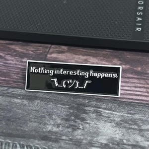 May include: A black enamel pin with the text "Nothing interesting happens:" and a shrug emoticon. The pin has a white border and is rectangular. The pin is on a textured surface.