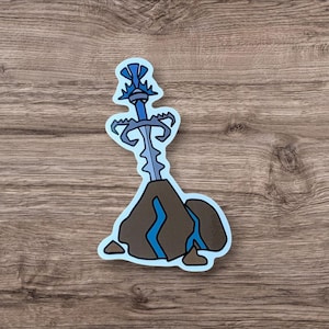 May include: A sticker featuring a blue sword embedded in a brown rock formation with blue accents. The sword has a detailed hilt and blade. The sticker has a light blue border and is set against a wood grain background.