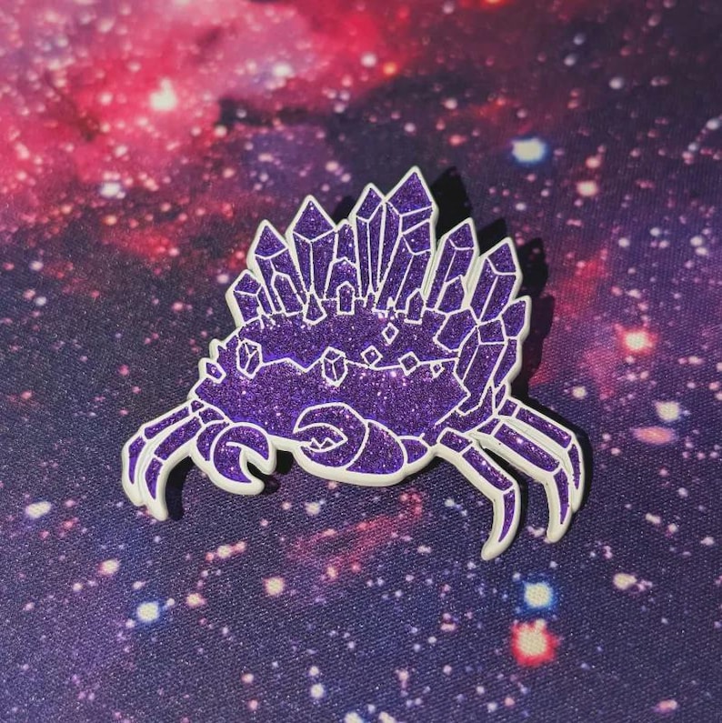 K&ouml;nnte beinhalten: Ein lila glitzernder Emaille-Pin in Form einer Krabbe mit Kristallformationen. Die Krabbe hat wei&szlig;e Umrisse und ist vor einem Hintergrund im Galaxien-Design platziert.