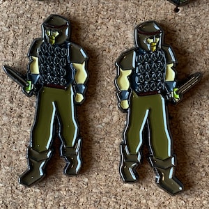 May include: Two metal pins depicting a fantasy warrior figure. The figures are wearing chainmail armor, helmets, and holding swords. The figures are green, black, and gold.