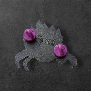 K&ouml;nnte beinhalten: Nahaufnahme der R&uuml;ckseite eines Pins mit zwei lila Ansteckern. Der Pin ist dunkelgrau mit einem stilisierten Design und dem Text "Becks". Der Pin liegt auf einer dunkelgrauen Oberfl&auml;che.