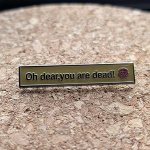 May include: A rectangular enamel pin with the text "Oh dear, you are dead!" in black lettering on a gold background. A red and gold emblem is on the right side. The pin is set against a textured, brown surface.