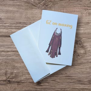 May include: A light blue greeting card with the text "GZ on maxing" in gold lettering. The card features a maroon and tan illustration of a cape. A matching light blue envelope is next to the card.
