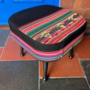 May include: A square footstool with four black tapered legs. The top and sides feature a black fabric base accented with a wide, horizontal band of woven textile in vibrant stripes of green, pink, yellow, and blue, including bird motifs.