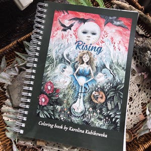 Peut inclure: Un livre de coloriage à reliure spirale intitulé Rising par Karolina Kubikowska. La couverture présente une illustration détaillée d'une personne avec un troisième œil, une grande lune, des corbeaux et des éléments forestiers en rouge, bleu, vert et blanc.