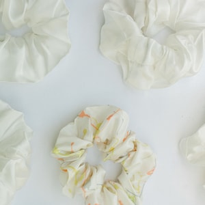 May include: Five white scrunchies with a variety of patterns. One scrunchie has a white background with orange, yellow, and green paint splatters. The other four scrunchies are plain white.