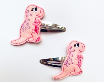 May include: Two pink dinosaur hair clips with embroidered details. The dinosaurs are facing right and have black eyes. The clips are silver.