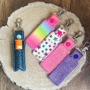 Könnte beinhalten: Fünf bunte Schlüsselanhänger mit Glitzer und verschiedenen Mustern. Jeder Schlüsselanhänger hat eine Schlaufe zum Anbringen von Schlüsseln und eine kleine Tasche zum Aufbewahren von Lippenbalsam. Die Schlüsselanhänger sind in den Farben Rosa, Lila, Blau, Weiß und Regenbogen.