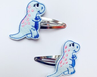 May include: Two light blue dinosaur hair clips with pink and blue embroidered details. The dinosaurs are standing on their hind legs with their front legs raised. The clips are silver.
