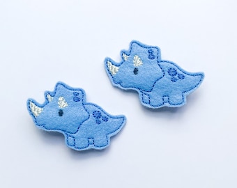 May include: Two blue felt dinosaur appliques with silver stitching. The dinosaurs are in a triceratops shape with three horns and a frill.