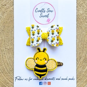 May include: Two hair accessories: a bow and a bee clip. The bow has a white background with yellow bees and glittery yellow sections. The bee clip is yellow and black with a smiling face. The card reads "Crafts Sew Sweet".
