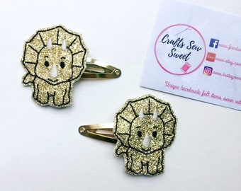 May include: Two gold glitter felt dinosaur hair clips with white stitching. The dinosaurs are facing forward with their mouths closed. The clips are attached to gold metal alligator clips.
