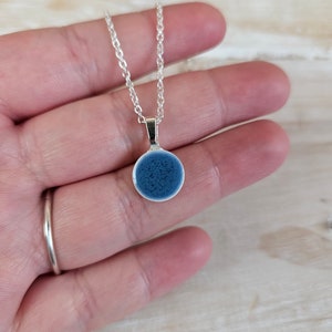 May include: A silver necklace with a small, circular pendant. The pendant has a dark blue, textured pattern. The necklace is suspended from a fine silver chain. The background is a neutral tone.