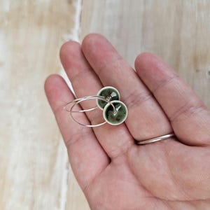 May include: Silver hoop earrings featuring two round, dark green and white ceramic beads. The earrings are held in a hand, with a silver ring on the finger. The background is a light wood surface.