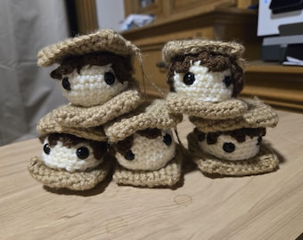 Könnte beinhalten: Ein Stapel gehäkelter Marshmallows, jeder mit einem braunen Häkelhut und einem weißen Häkelgesicht mit schwarzen Augen. Die Marshmallows sind auf einer Holzoberfläche gestapelt.