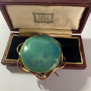 May include: A green and blue oval cameo brooch with a gold setting. The brooch is in a red velvet box with a label that reads "R.A.C.S. Jewellers, London, S.E."