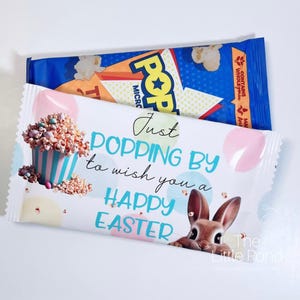 Può includere: Una borsa regalo bianca e blu con un coniglio e popcorn. La borsa ha il testo "Just Popping By to wish you a Happy Easter".