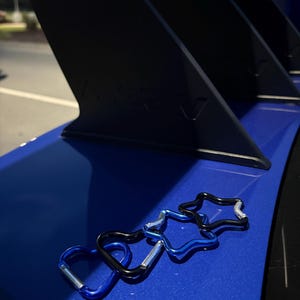 Puede incluir: Cuatro peque&ntilde;os mosquetones con formas de corazones y estrellas, con acabados en azul met&aacute;lico y negro y cierres de resorte plateados, sobre una superficie azul lisa y brillante frente a un componente de aler&oacute;n de autom&oacute;vil negro.