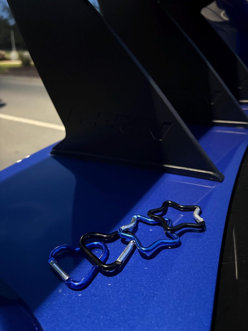 Puede incluir: Cuatro peque&ntilde;os mosquetones con formas de corazones y estrellas, con acabados en azul met&aacute;lico y negro y cierres de resorte plateados, sobre una superficie azul lisa y brillante frente a un componente de aler&oacute;n de autom&oacute;vil negro.