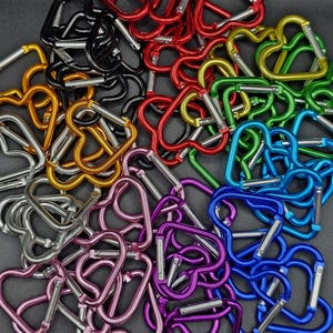 May include: A collection of heart-shaped carabiners in various metallic colors including red, black, gold, silver, green, blue, and purple. Each carabiner features a spring-loaded gate mechanism for opening and closing, arranged in a dense pile.
