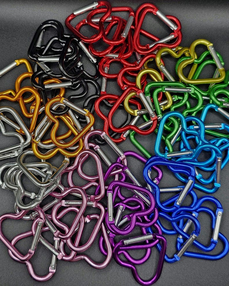 May include: A collection of heart-shaped carabiners in various metallic colors including red, black, gold, silver, green, blue, and purple. Each carabiner features a spring-loaded gate mechanism for opening and closing, arranged in a dense pile.