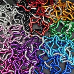 Puede incluir: Una colecci&oacute;n de mosquetones con forma de estrella en varios colores met&aacute;licos, incluyendo rojo, dorado, verde, azul, morado, rosa, plateado y negro. Cada mosquet&oacute;n cuenta con un mecanismo de cierre de resorte, dispuestos en un mont&oacute;n.