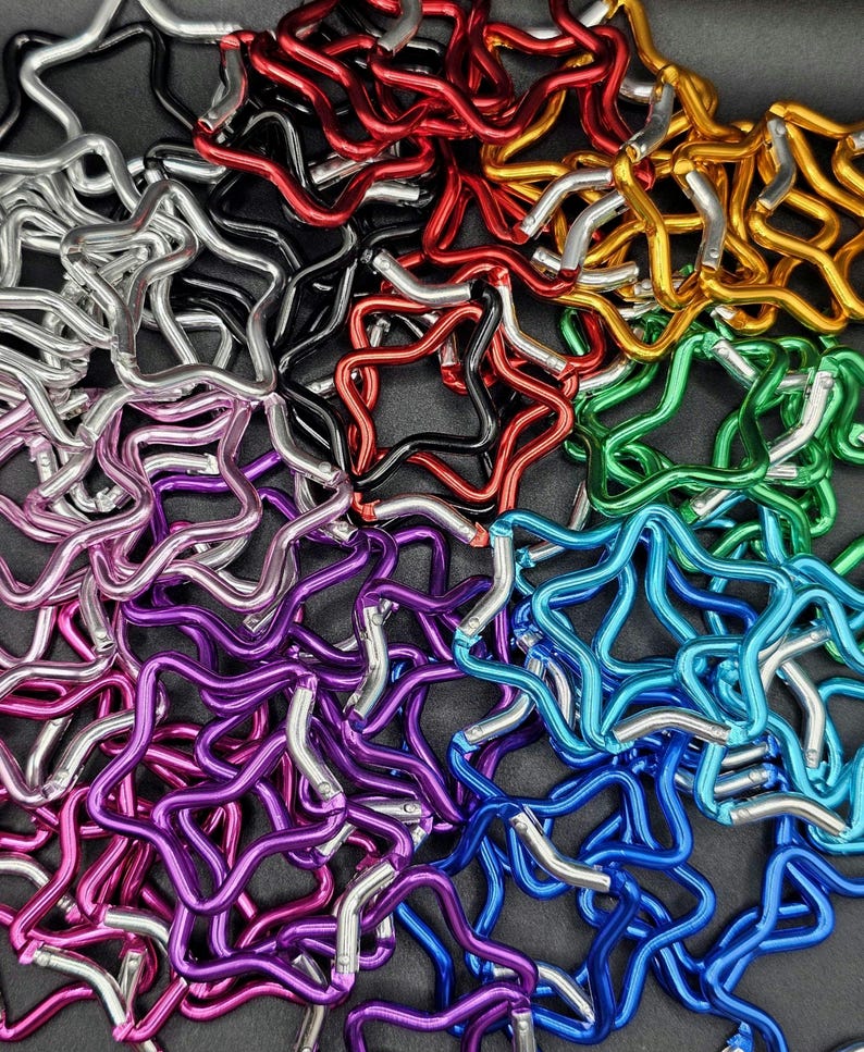Puede incluir: Una colecci&oacute;n de mosquetones con forma de estrella en varios colores met&aacute;licos, incluyendo rojo, dorado, verde, azul, morado, rosa, plateado y negro. Cada mosquet&oacute;n cuenta con un mecanismo de cierre de resorte, dispuestos en un mont&oacute;n.