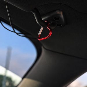 May include: A small, heart-shaped red metal carabiner clip attached to a black plastic hook mounted on the interior ceiling of a vehicle. The carabiner features a spring-loaded silver gate mechanism and a smooth, glossy finish on the red frame.