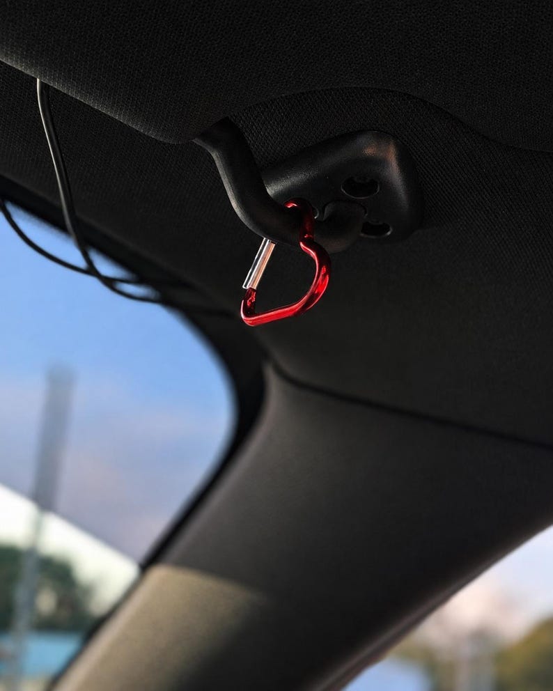 May include: A small, heart-shaped red metal carabiner clip attached to a black plastic hook mounted on the interior ceiling of a vehicle. The carabiner features a spring-loaded silver gate mechanism and a smooth, glossy finish on the red frame.