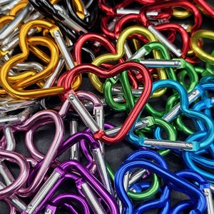 May include: A collection of heart-shaped carabiners in various metallic colors including red, blue, green, gold, purple, pink, and black. Each carabiner features a spring-loaded silver gate mechanism for opening and closing the heart-shaped frame.