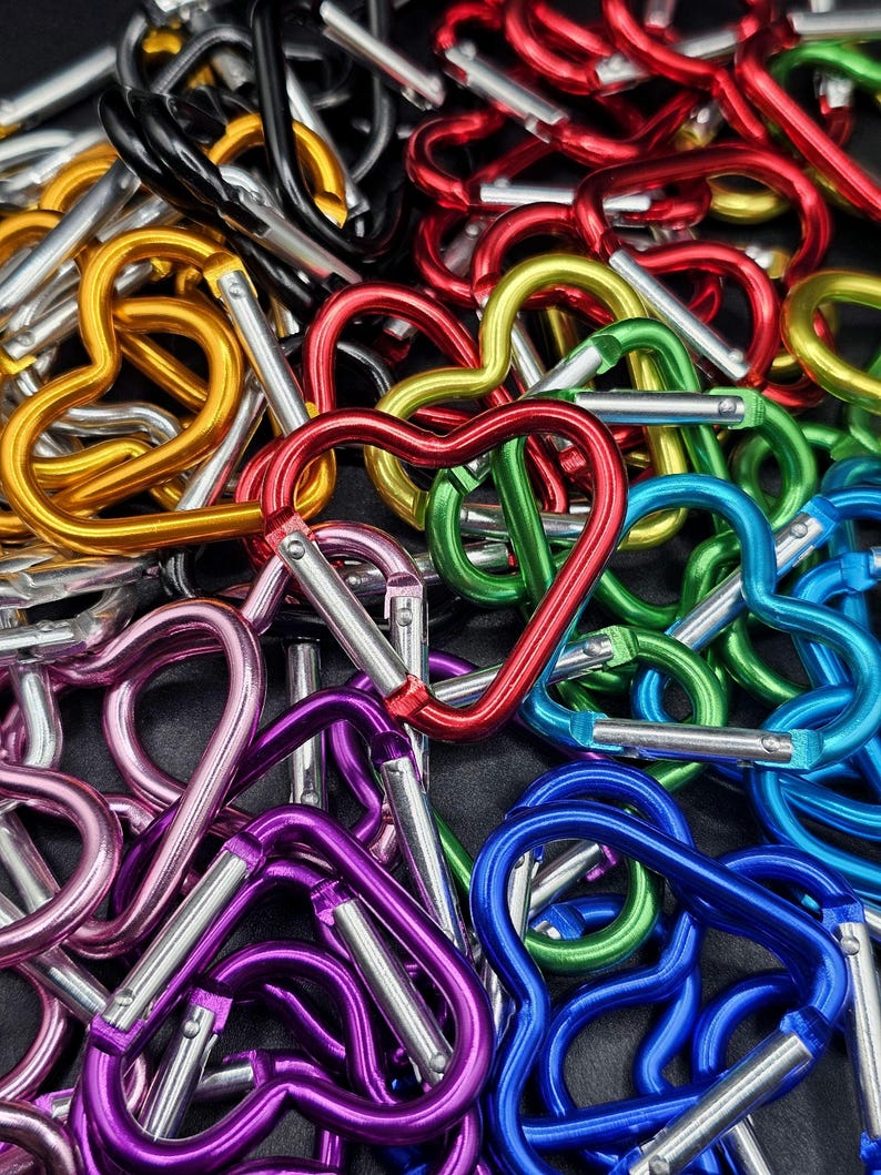 May include: A collection of heart-shaped carabiners in various metallic colors including red, blue, green, gold, purple, pink, and black. Each carabiner features a spring-loaded silver gate mechanism for opening and closing the heart-shaped frame.